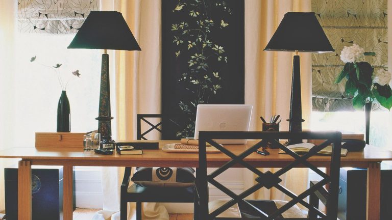 Oubliez la caféine – Les aromathérapeutes utilisent cette huile essentielle comme un moyen rapide et naturel de réinitialiser instantanément la concentration et de soulager la fatigue mentale. Home office space with black lamps, black chairs, and a large wooden desk with a laptop and pens. To either side are bright windows with patterned blinds, flowers, and vases.
