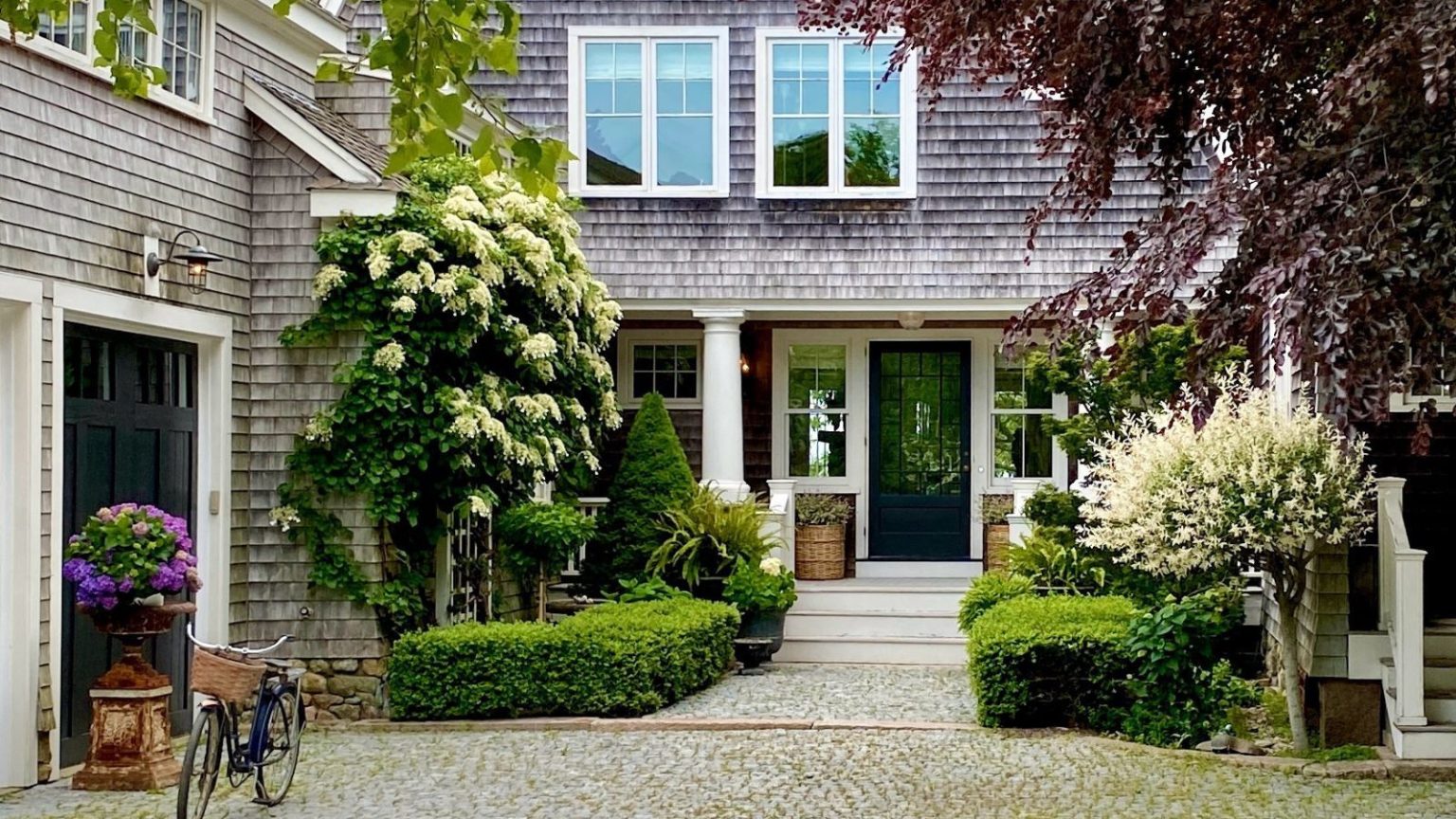 20 décorations de porche printanières pour rafraîchir votre entrée pour « la plus jolie saison de l’année » Shingle style house with front porch with planters, hedges, and two garages with a bicycle outside