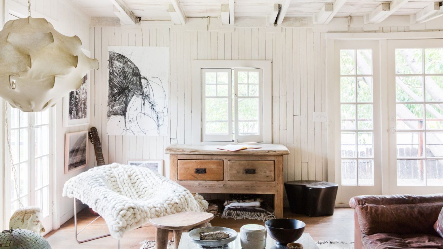 Les couleurs vives pourraient être au sommet des tendances en matière de design, mais Leanne Ford affirme que les tons neutres ne vont nulle part – voici comment la créatrice introduit les classiques en 2026 Living room with white shiplap walls, French doors, white beamed ceiling, wooden floor, a wooden console underneath the window, a metal armchair with a chunky knit throw on top and a leather sofa