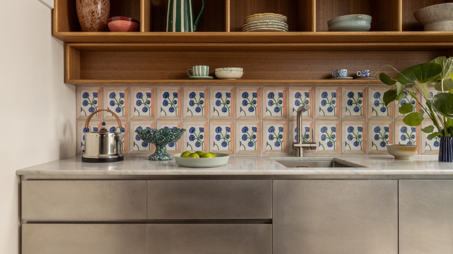 Les designers utilisent ce mélange de matériaux haut-bas pour créer les cuisines les plus chics de 2026 – c’est un moyen garanti d’adopter la tendance minimaliste douce A modern kitchen counter featuring brushed stainless steel cabinets and a white marble countertop. The backsplash consists of decorative hand-painted floral tiles, set beneath warm wood open shelving filled with ceramic bowls and glassware.