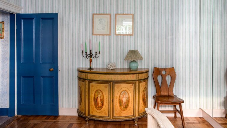 3 choses pour lesquelles les antiquaires disent toujours que vous devriez faire des folies – et 3 choses pour lesquelles ils ne gaspillent jamais d’argent Bedroom with a blue painted door, striped blue wallpaper, a vintage console table with a painted design, an iron candelabra on top with colourful candles, and a vintage chair beside it
