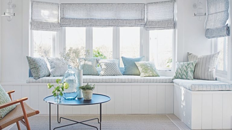 A bright bay window seating area with large windows, sheer grey blinds, patterned cushions, and a circular coffee table with flowers and a large vase, with a wooden rattan chair.