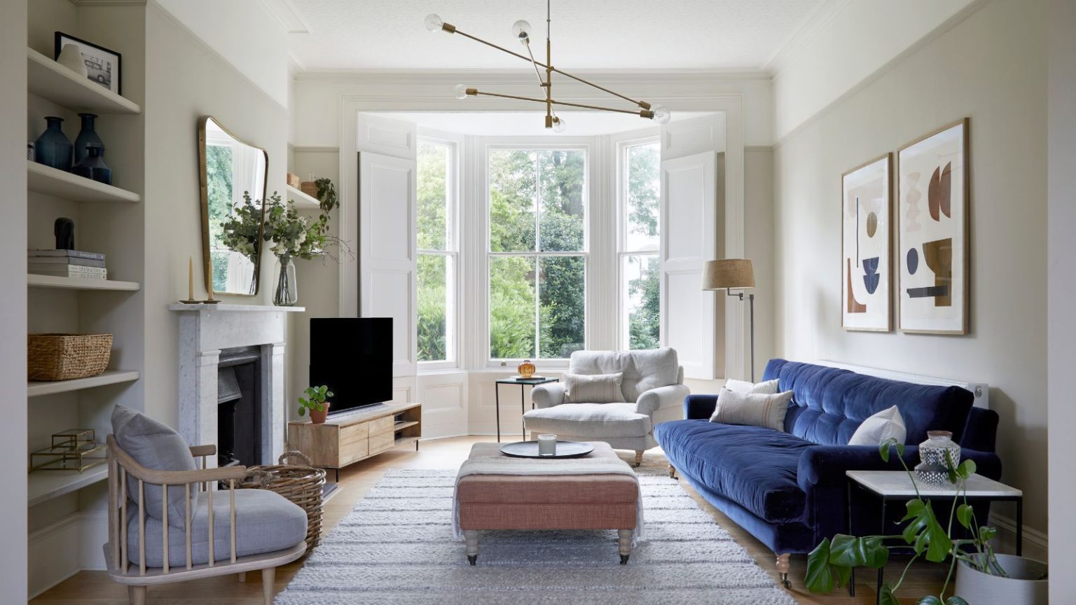 Les 4 astuces de mise en page intelligentes utilisées par les concepteurs pour rendre un téléviseur moins dominant dans votre salon Living room with taupe walls, large bay window, grey rug at the centre, a pink ottoman on top, dark blue velvet sofa, and a large fireplace with a mirror above it