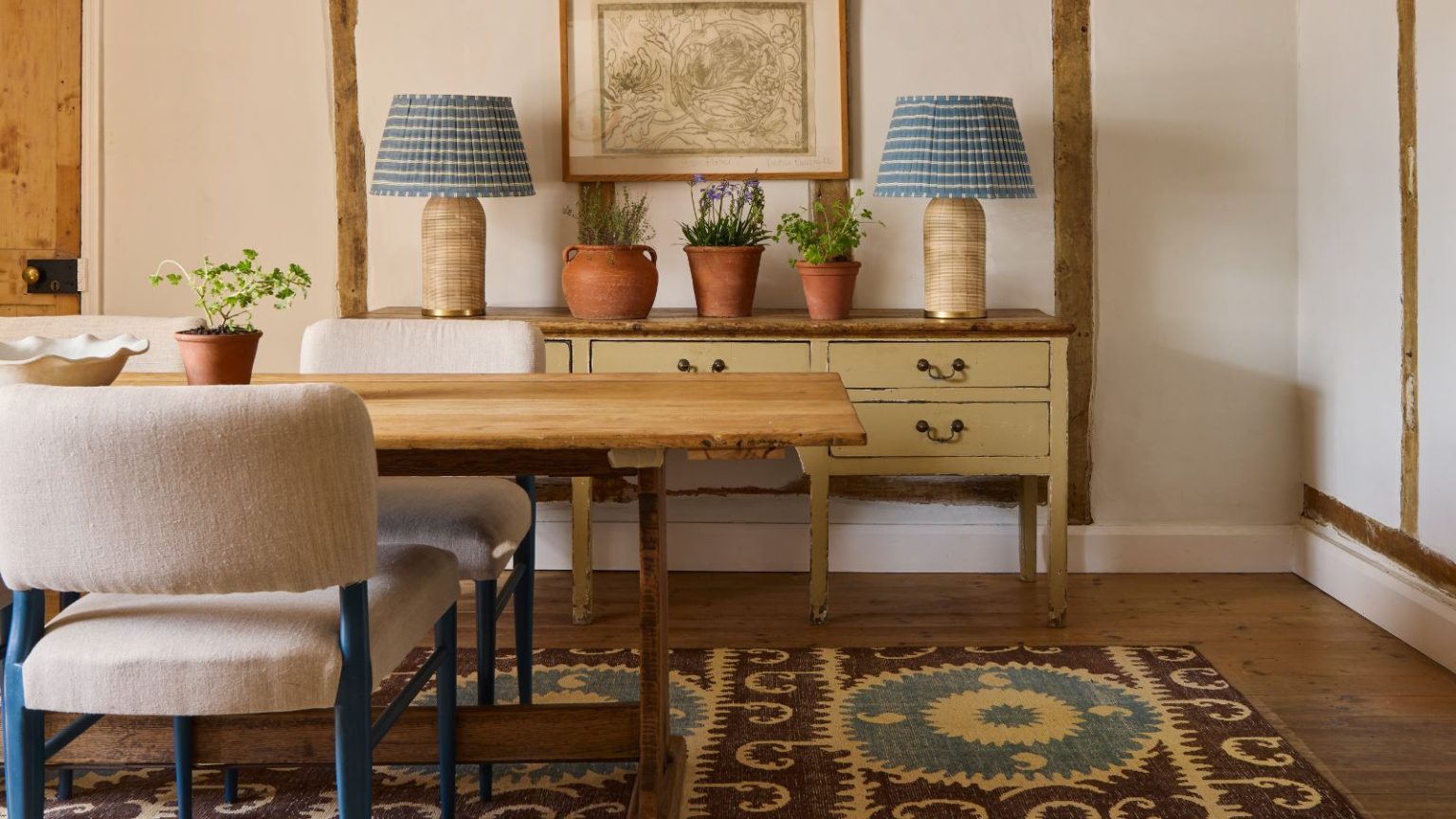 Ce sont les 4 couleurs de tapis à éviter en 2026 – et les teintes que les designers utilisent à la place Dining room with a wooden painted console with two large gingham blue lamps on top, a wooden long dining table, upholstered dining chairs with a blue frame, and a brown and blue kilim rug beneath