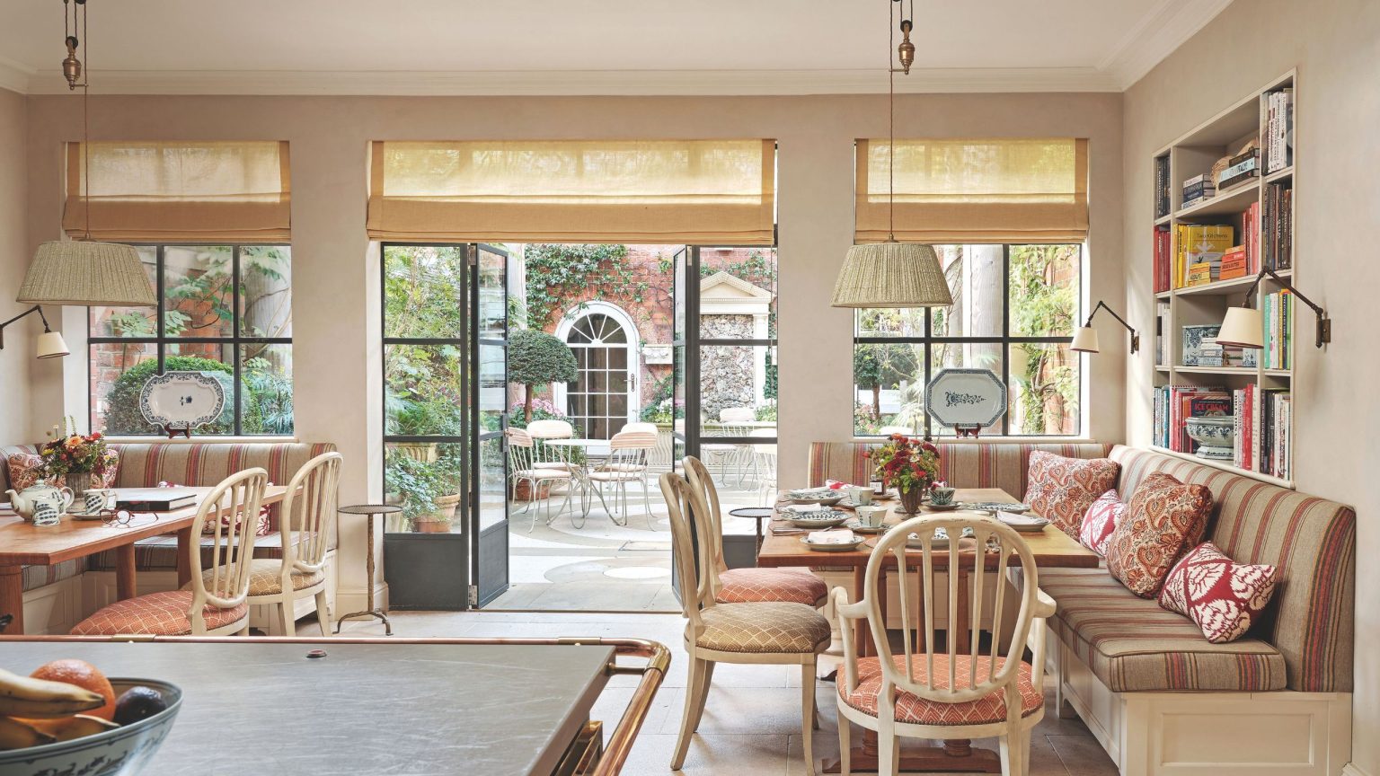 Comment une maison de campagne anglaise historique a été transformée en une maison familiale habitée qui privilégie l’authenticité et le confort A bright breakfast nook with built-in banquette seating, patterned pillows, and large glass doors that open onto a lush garden courtyard