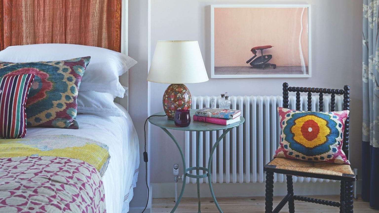 Votre linge de lit est-il moins frais au printemps ? Des experts en blanchisserie révèlent la routine de lavage idéale pour les mois les plus chauds A colorful bedroom with a white bed with colorful cushions and blankets, a black chair, a clear circular table with a colorful lamp with white shade, and a large artwork hung above the radiator.