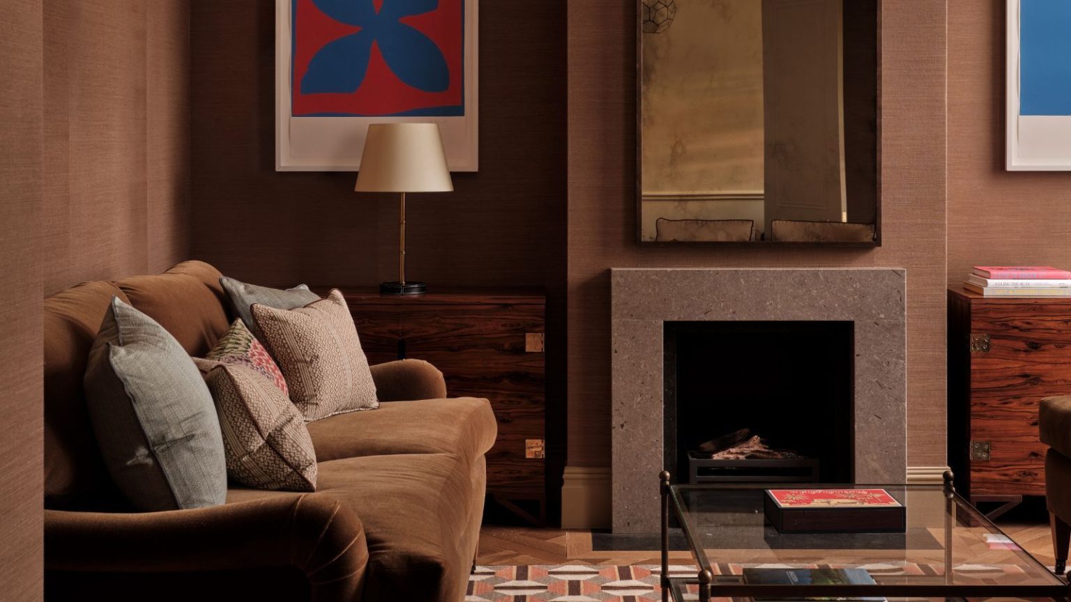 Pourquoi « Hue Drenching » est le secret du concepteur pour rendre les petites pièces plus intéressantes – « C’est nuancé et réfléchi » Living room with brown grasscloth walls, cream ceiling, lantern pendant light, geometric orange and brown rug, glass coffee table, brown velvet sofa, and a fireplace with tarnished mirror above it