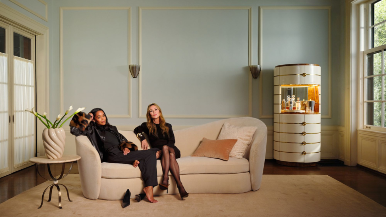 a large living room with two women sitting on a cream couch, a large bar cabinet to the left, and a side table to the right with a vase of flowers on it