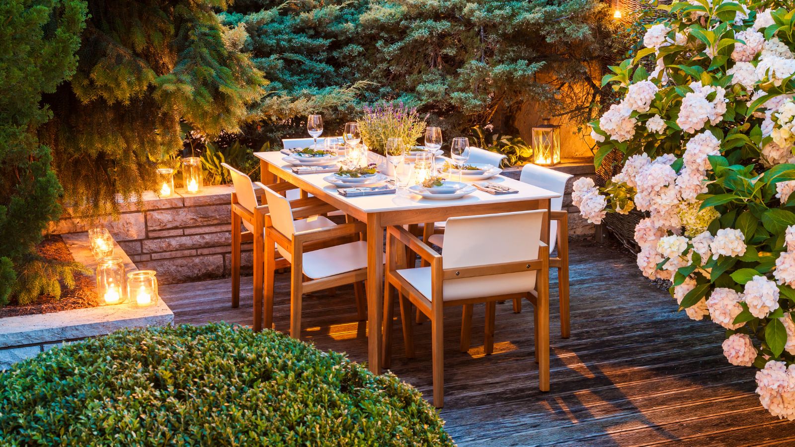 Patio avec table à manger en bois pour accueillir le dîner, entouré de verdure et éclairé par des bougies allumées