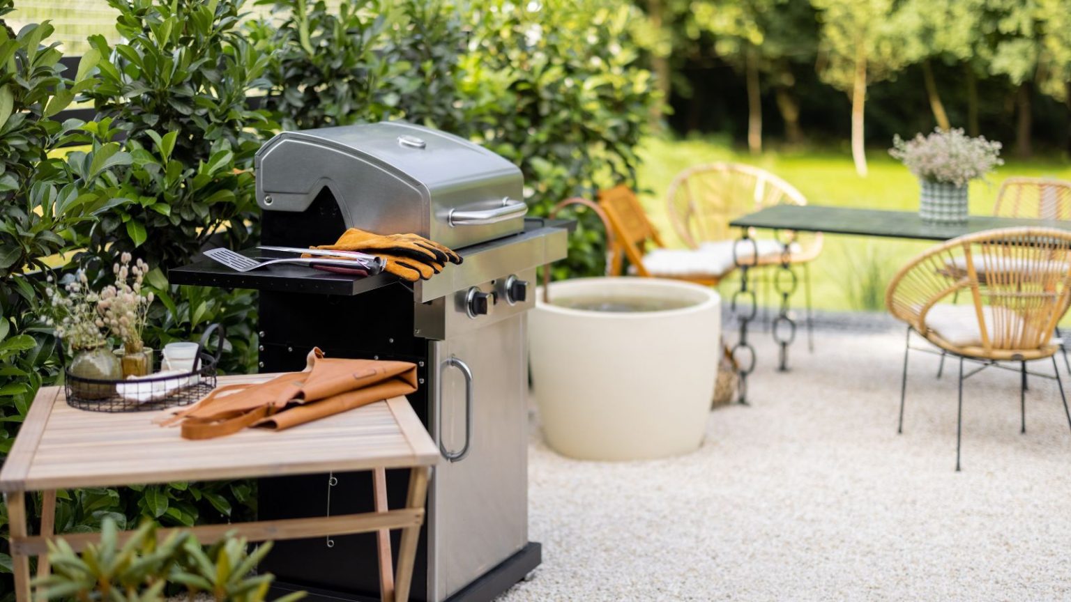 A freestanding steel grill against a hedge line in a backyard. A wooden table with an apron on it beside it.