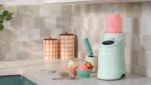 A small light blue ice cream maker on a kitchen counter surrounded by small bowls of ice cream. The clear churning compartment is filled with pink mixture.