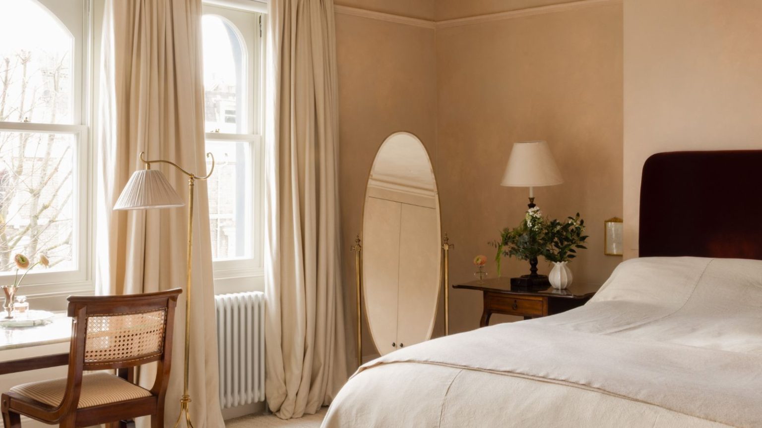 Avez-vous entendu parler de « Peige » ? Ce sont les designers neutres, doux et apaisants qu’adoptent réellement en 2026 Bedroom with plaster pink walls, dark red velvet bed, linen bedding, round floor mirror, vintage desk with rattan chair, floor lamp and large arched windows