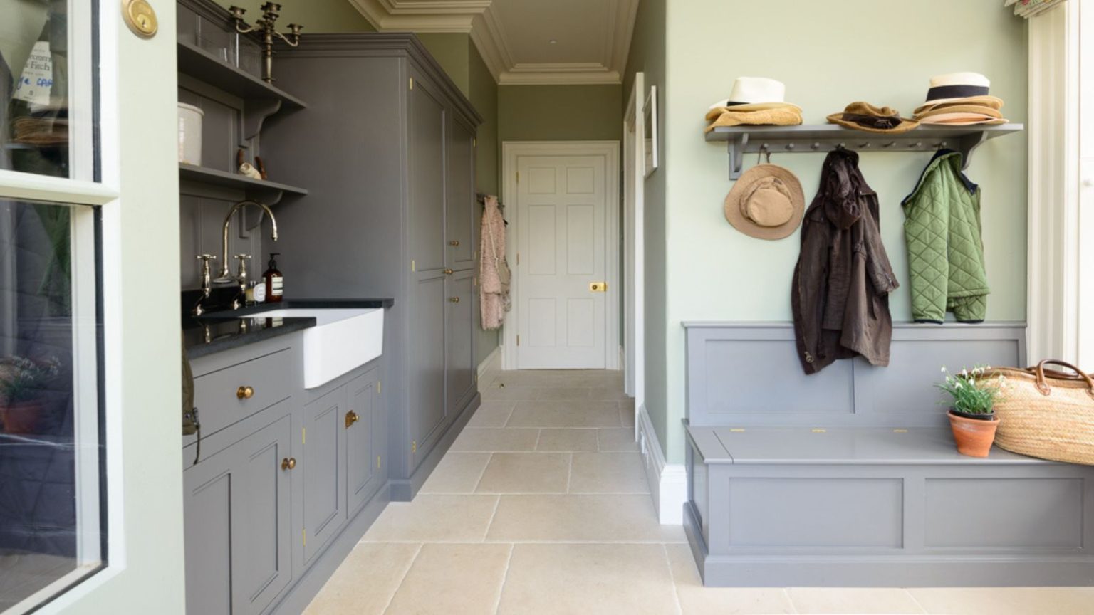 Enfin, plus de vadrouille bruyante qui envahit ma cuisine – Cette solution de stockage simple à 12 $ a instantanément rendu tout plus propre, plus propre et beaucoup plus organisé. Pastel green utility room and mudroom gray cabinets, storage bench and butler sink. It has cream stone floors and mounted shelves and hooks for added storage.