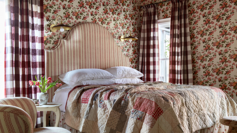 a heritage maximalist bedroom with cherry printed wallpaper, a striped headboard, a plaid quilt, checker printed drapes, and four pillows on the bed