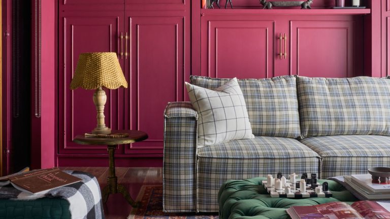 Living room with raspberry pink cabinetry, plaid blue sectional sofa, Persian rug, green velvet ottoman, side table with lamp and plant on the floor