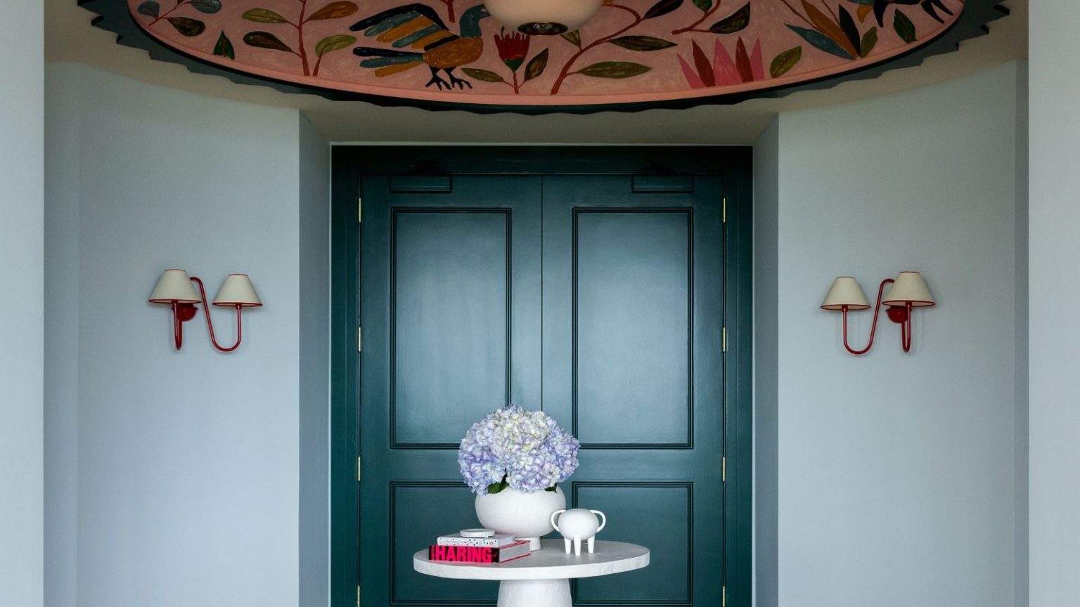 Cette entrée utilise la lumière empruntée par Farrow & Ball pour prouver que les couleurs vives peuvent être à la fois calmes et dramatiques. Pale blue entryway with dark teal double doors, two red sconces on walls, a large pink floral ceiling mural, a round, small white table in the centre with a vase and books, and parquet wooden floors
