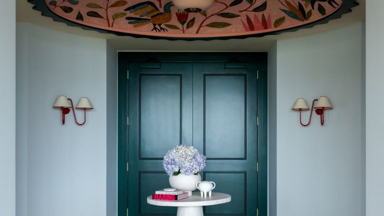 Cette entrée utilise la lumière empruntée par Farrow & Ball pour prouver que les couleurs vives peuvent être à la fois calmes et dramatiques. Pale blue entryway with dark teal double doors, two red sconces on walls, a large pink floral ceiling mural, a round, small white table in the centre with a vase and books, and parquet wooden floors