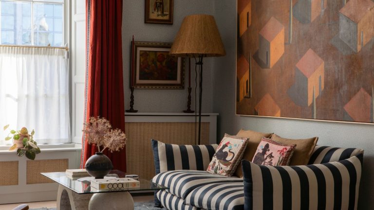 Living room with pale blue walls, striped sofa, red curtains, a glass and stone coffee table, a large painting above the sofa in orange and red of houses, and a rattan floor lamp in the corner