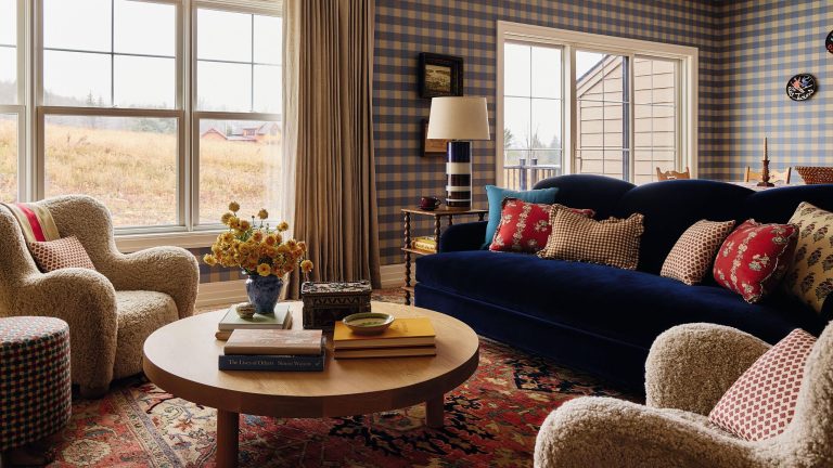 an open plan living area in a home in the Catskills with blue gingham wallpaper, a navy velvet couch, vintage red rug, sheepskin armchairs and a round wooden coffee table