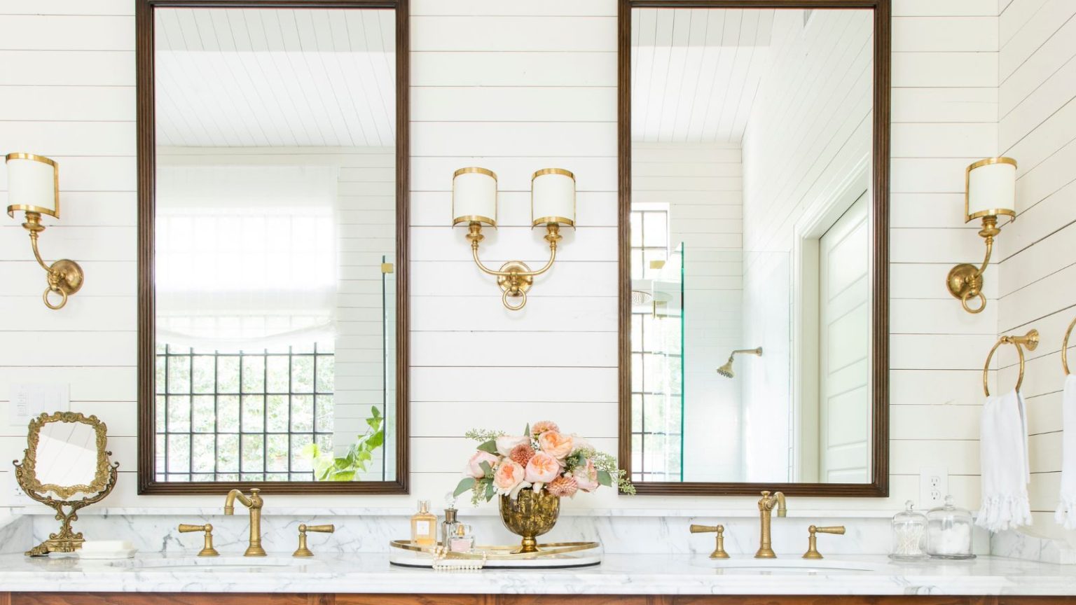 Bathroom with Cloud White painted panelled walls, large mirrors with wooden frames, a marble vanity unit with double sinks and gold taps, and gold and white sconces flanking the mirrors