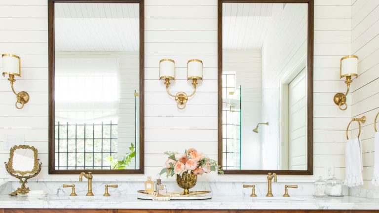 Bathroom with Cloud White painted panelled walls, large mirrors with wooden frames, a marble vanity unit with double sinks and gold taps, and gold and white sconces flanking the mirrors