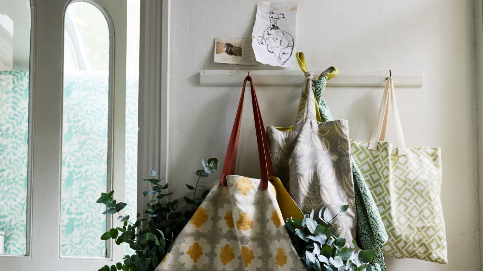 White wall with white wall hooks, with patterned tote bags hanging up. One is filled with eucalyptus. Above the shelf are two hand-drawn artworks, and to the left is an open white door.