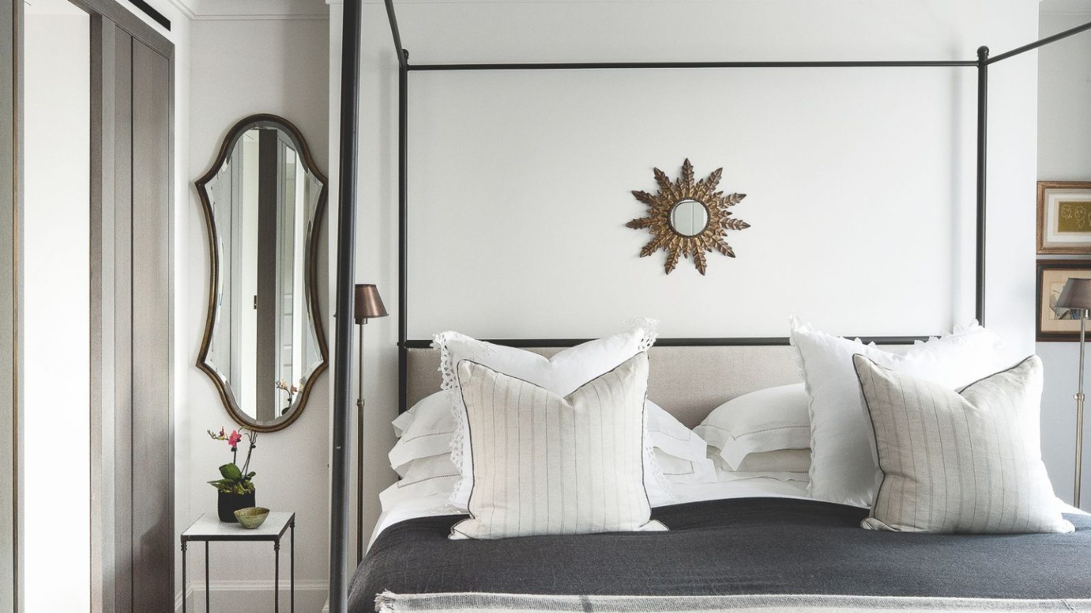 White bedroom with a black four-poster bed, grey bedding with white cushions, and a small circular mirror above the bed. To the left is an oval shaped mirror.