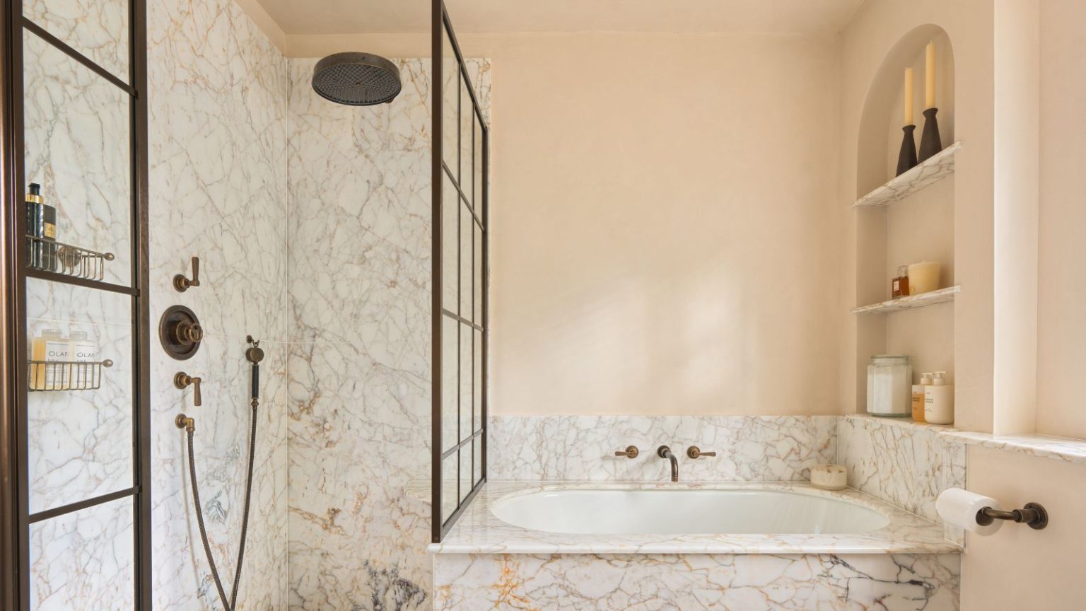 A small bathroom with marble tiles across the bath and shower, a recessed wall niche holding products and glass canisters, and black accent details