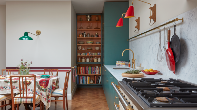 a kitchen with a small dining table, a long countertop with a pot rack hanging on the wall above, and a bookshelf in the background holding bowls, spices, and decor