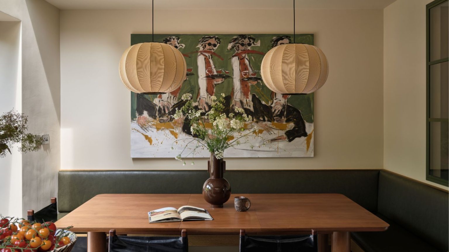 5 choix de conception qui affectent discrètement votre humeur et votre bien-être – et comment y remédier pour l’été Dining nook with banquette green leather seating, a wooden table, a pair of lantern lights hanging above, an abstract painting of people dancing behind, and a large floral arrangement in a brown vase