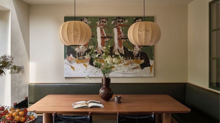 5 choix de conception qui affectent discrètement votre humeur et votre bien-être – et comment y remédier pour l’été Dining nook with banquette green leather seating, a wooden table, a pair of lantern lights hanging above, an abstract painting of people dancing behind, and a large floral arrangement in a brown vase