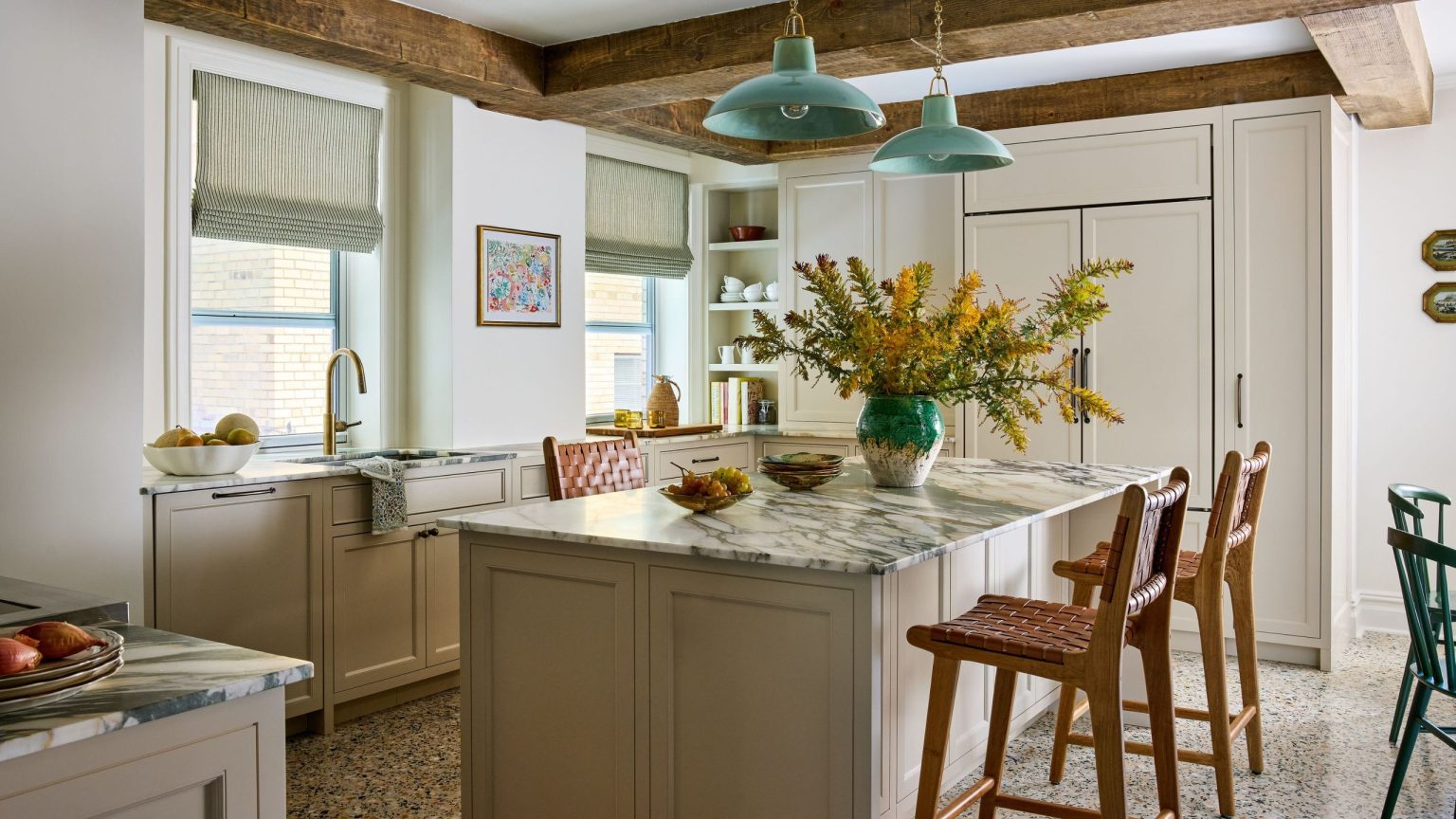 A kitchen with neutral cabinets, white walls, marble counters and a kitchen island with two tan bar stools and wood ceiling beams.