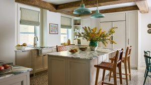 Cette cuisine d’appartement de New York est remplie de solutions de rangement cachées et intelligentes – et elle est aussi élégante que fonctionnelle A kitchen with neutral cabinets, white walls, marble counters and a kitchen island with two tan bar stools and wood ceiling beams.