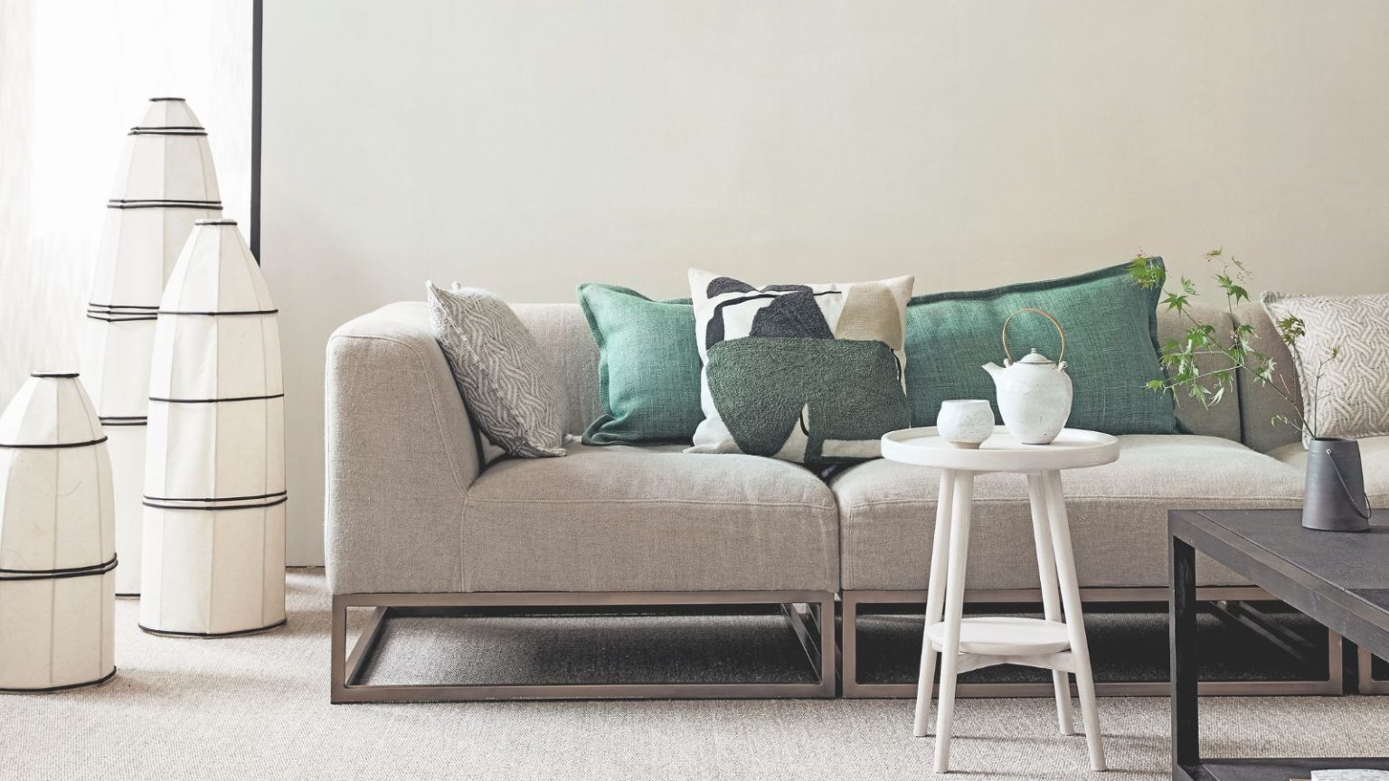 A white living room with a light grey couch, green cushions, a white table with white tea set, black coffee table with green plant, and three black and white large lamps beside the couch.