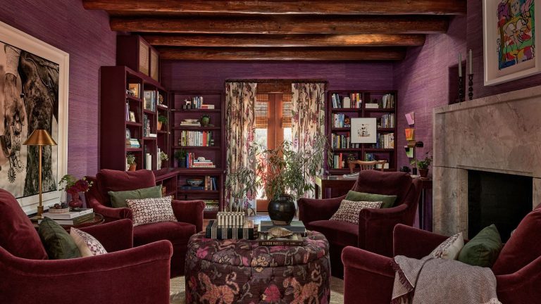 4 couleurs vintage que les créateurs jurent faire leur retour en 2026 Living room with deep purple grasscloth walls, beamed ceiling, floral drapes covering glass doors, burgundy bookshelves, four maroon velvet armchairs sat around a floral ottoman with decor on top