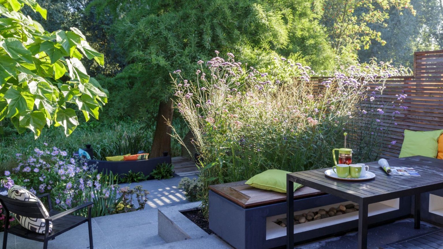 Patio seating area in sunny garden with a table, bench seating, cafetiere, coffee cups, and colorful cushions. Beyond the patio is a black chair with a black and white cushion, flowers, and trees.