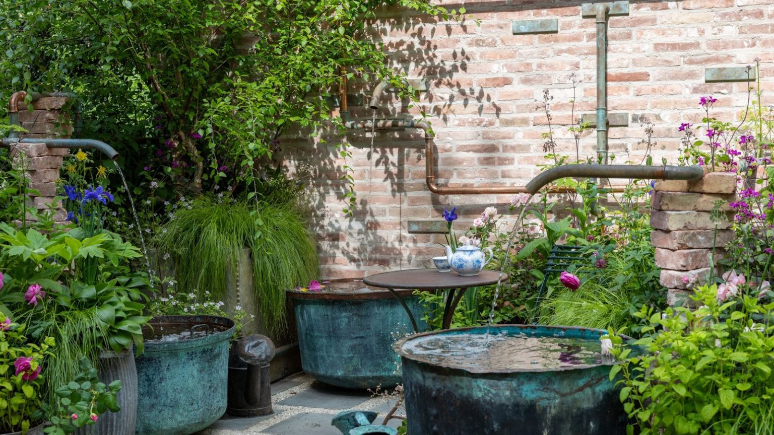 6 solutions de refroidissement pour la cour qui aident les plantes à vaincre la chaleur et à rester luxuriantes tout l’été Courtyard garden with water pipes flowing into large blue copper pots and lush, cottage-style planting