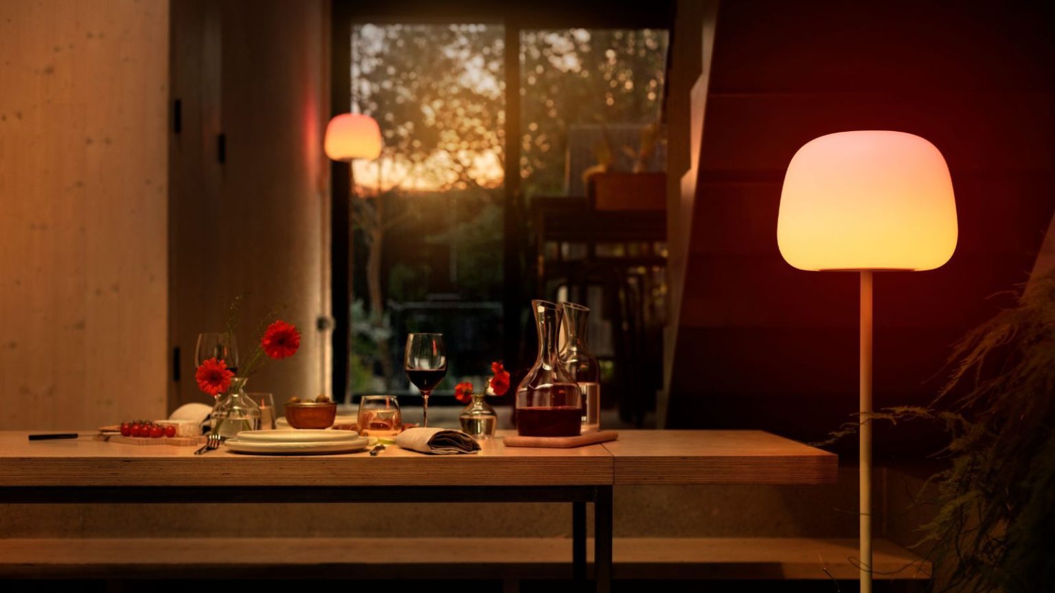 Govee smart floor lamp beside a long wooden dining table with tableware and a carafe of wine, with a view of the back garden