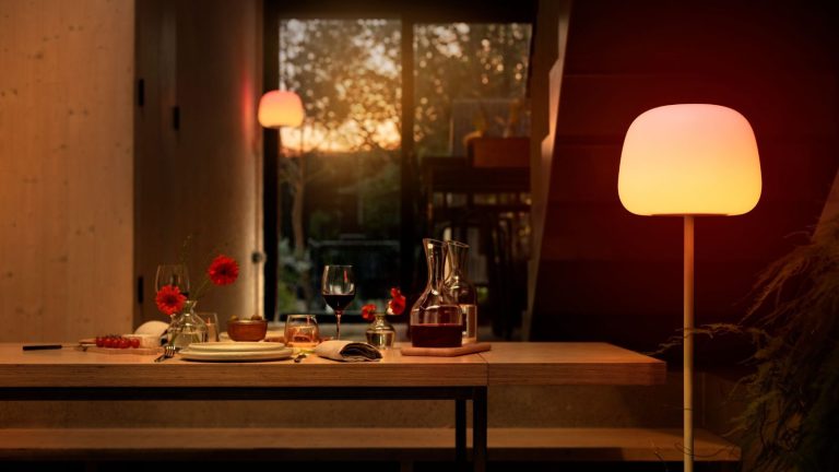 Réimaginez l’éclairage de votre maison avec une lampe pour chaque pièce et chaque instant Govee smart floor lamp beside a long wooden dining table with tableware and a carafe of wine, with a view of the back garden