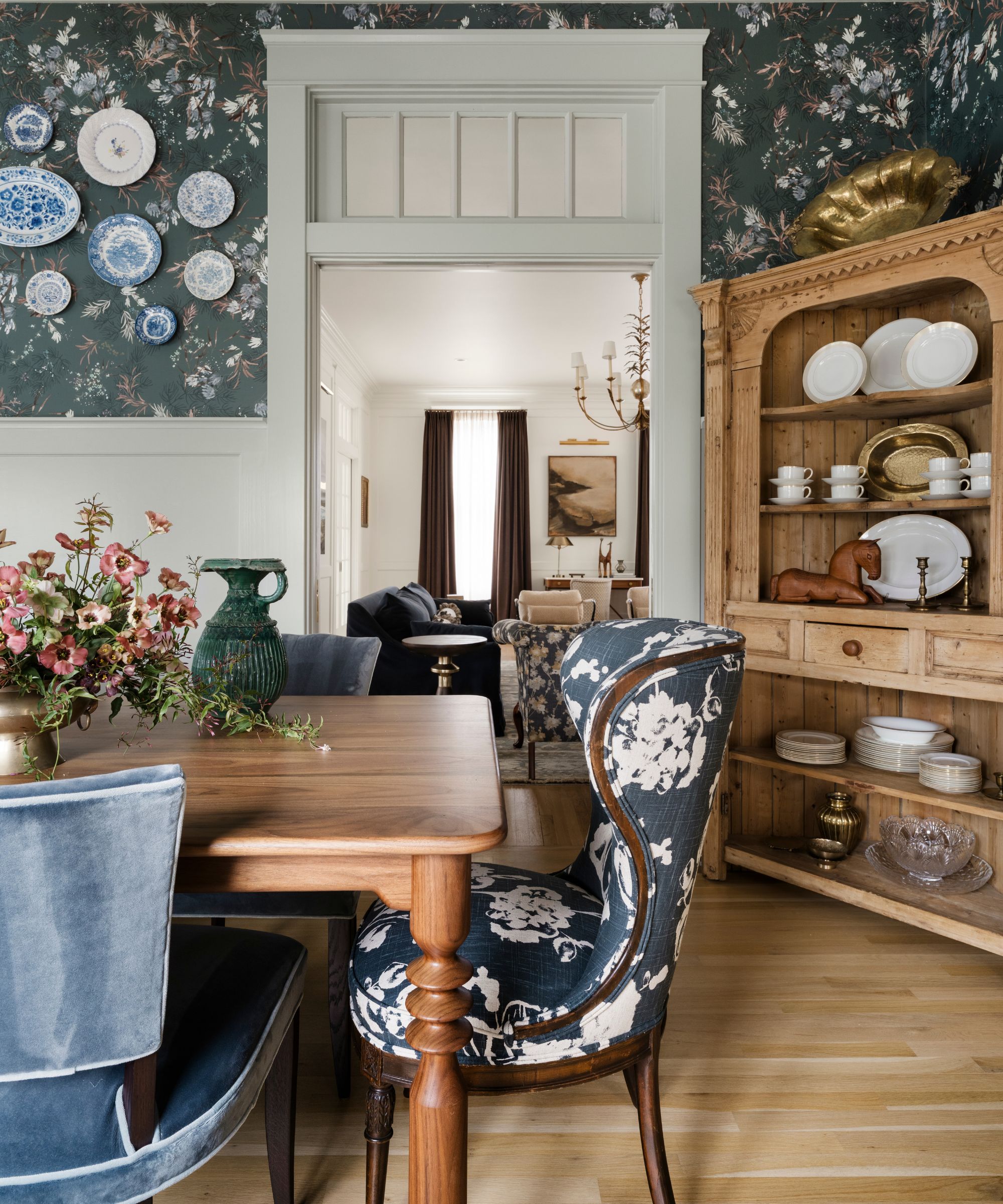 une salle à manger formelle avec un papier peint à fleurs bleu marine, des lambris et des menuiseries d'origine, une table à manger et un clapier en bois avec un mélange de chaises bleues à fleurs et en velours