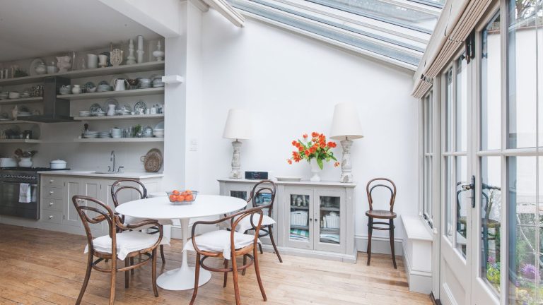 4 souvenirs de votre enfance que les concepteurs de maisons disent que vous devriez réellement conserver Conservatory with glass doors, round white breakfast table, wooden bistro chairs, a side console with two large lamps, leading into a kitchen with lots of open shelving decorated with ceramics