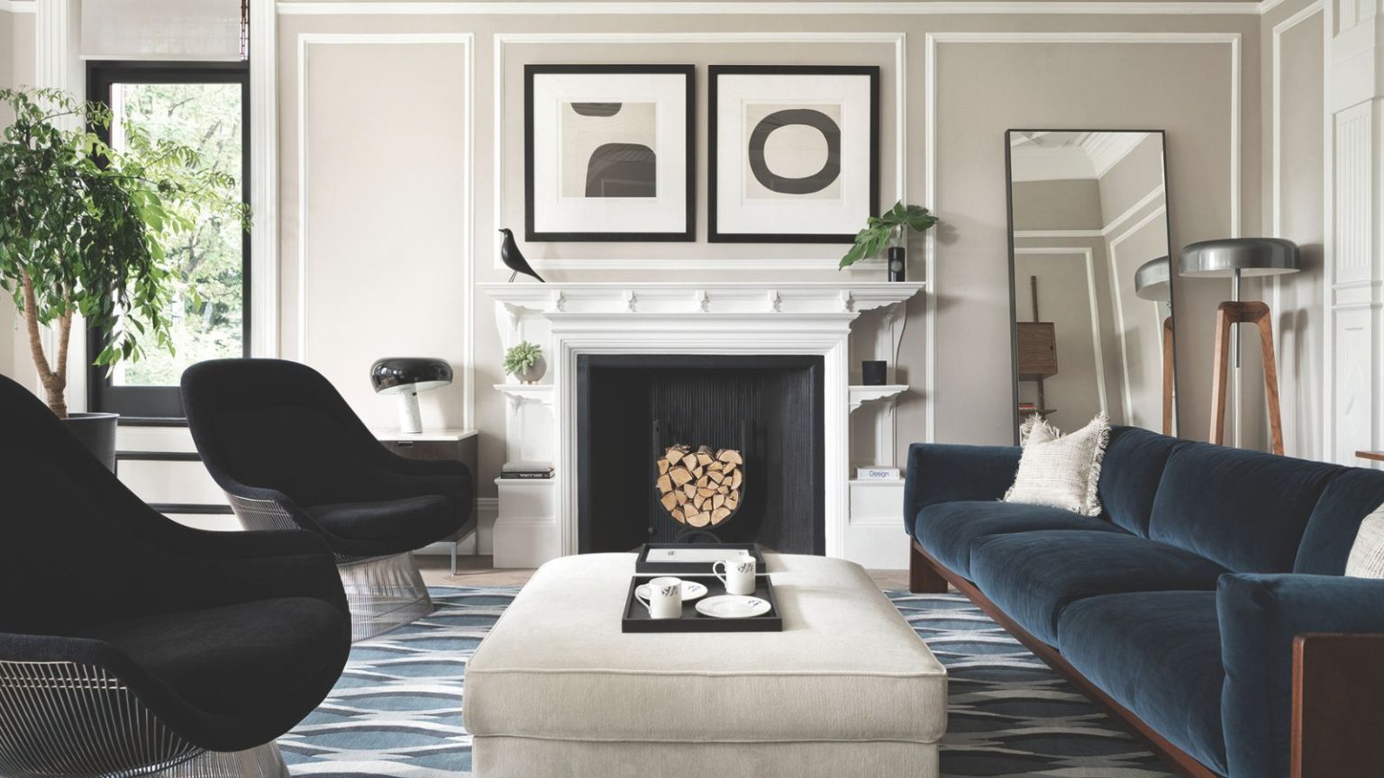 Ce sont les 3 couleurs que les designers disent de ne jamais associer avec du gris si vous voulez vraiment qu’il soit beau Living room with pale grey walls, blue velvet sofa, two black armchairs, a pale grey ottoman, a geometric blue rug, a large white fireplace and modern art hanging above it.