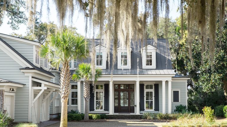 Le gris Boothbay de Benjamin Moore a peut-être 50 ans – mais les designers disent que c’est toujours une couleur simple et intemporelle pour 2026 Colonial-style home with Boothbay Gray painted exterior, large porch with dark brown front door, a white garage to the left, and a weeping willow and palm tree at the front of the home