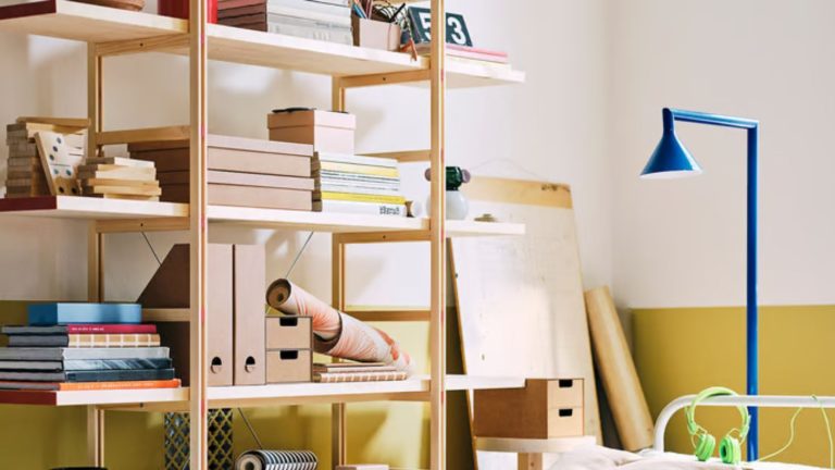40 ans plus tard, l’étagère la plus emblématique d’IKEA est de retour – et elle a subi une mise à jour en 2026, tout aussi chic mais tellement moins chère IKEA PS shelving unit decorated with books, files, and trinkets, against a yellow and white wall, with a blue floor lamp.