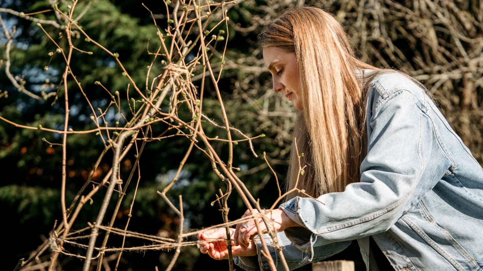 Woman in blue denim jacket and long hair placing natural jute twine around hazel wigwam