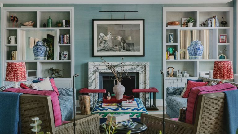 Living room with blue grasscloth walls, built-in bookshelves, marble fireplace with painting above it, pink armchairs with red lamps on either side, and two blue sofas.