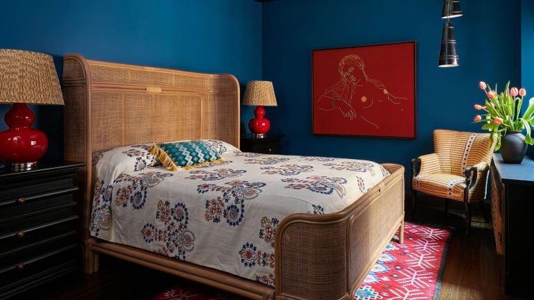 Déplacez-vous sur « Coastal Grandmother » – Les designers disent que cette nouvelle tendance élégamment éclectique la remplacera pour l’été 2026 Eccentric Aunt-style bedroom featuring teal walls, a rattan bed frame, abstract art work, a patterned accent chair, and mismatched bedding