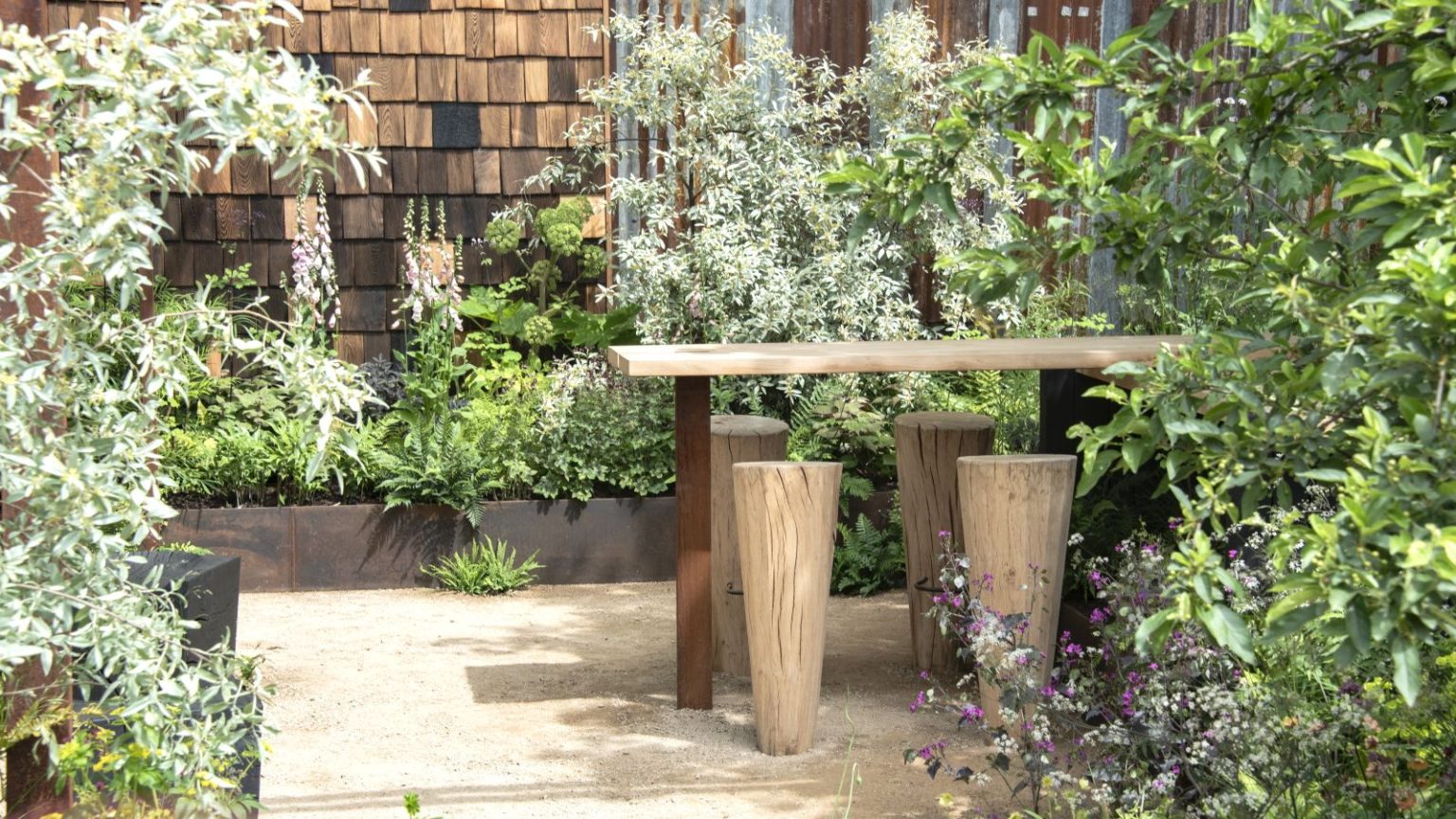 Cette erreur de jardinage facile à commettre en juin pourrait empêcher votre jardin de prospérer – voici ce qu’il faut faire à la place A wooden table and chairs surrounded by raised beds with green shrubs at the RHS Chelsea Flower Show 2026