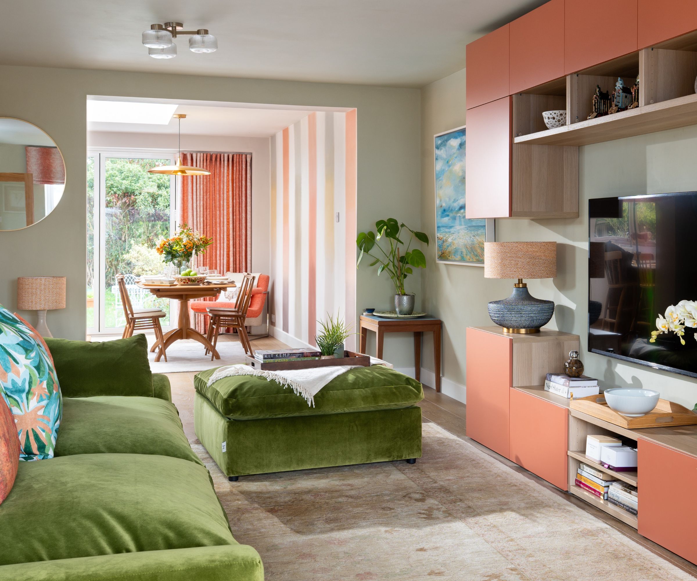Salon avec compartiments de rangement intégrés peints en Naperon par Farrow &amp; Ball, un canapé en velours vert et son pouf assorti, un tapis beige, une grande télévision, avec une salle à manger ouverte en arrière-plan.