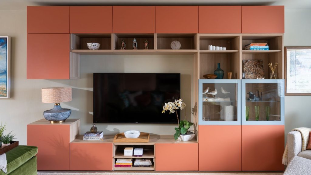 Living room with built-in cubby storage on the wall painted in Naperon by Farrow &amp; Ball, a large TV mounted on the wall at the centre, and a lamp and orchid on either side.